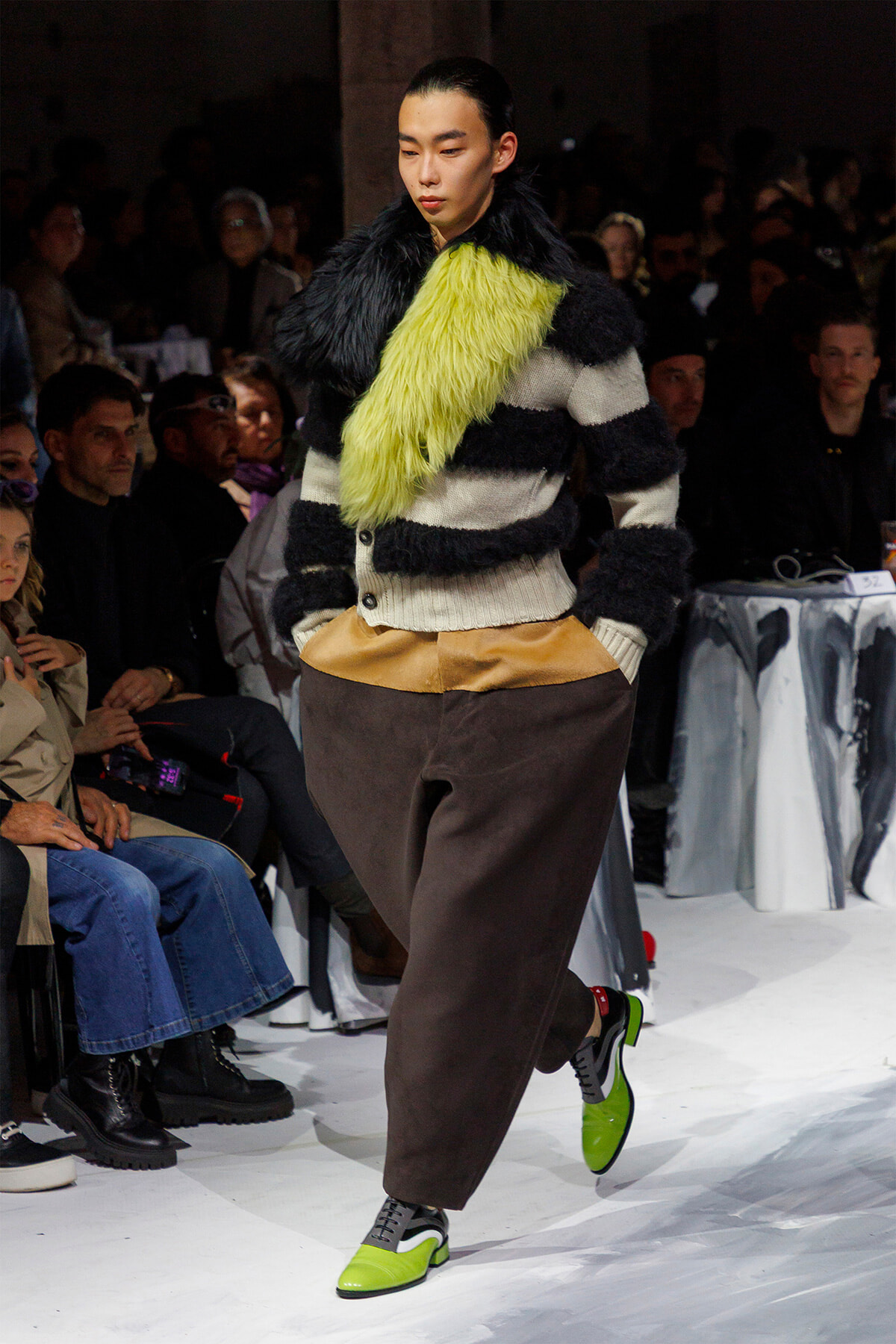 Model walks a fashion runway wearing a chunky striped fur cardigan, neon-yellow scarf, and brown wide-leg pants; audience seated nearby.