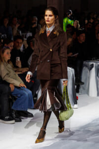 Model on a fashion runway wearing a brown double-breasted velvet blazer with a matching pleated midi skirt, carrying a pale green handbag.