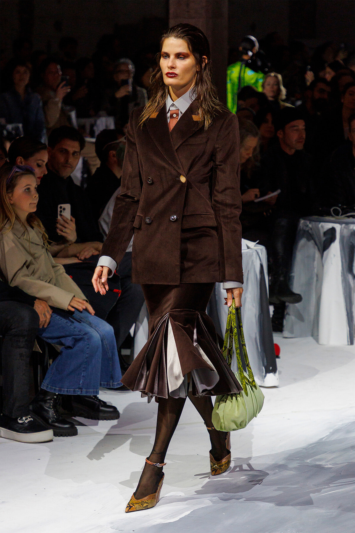 Model on a fashion runway wearing a brown double-breasted velvet blazer with a matching pleated midi skirt, carrying a pale green handbag.