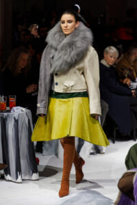 Model walks the runway in a cream leather jacket with an oversized gray fur collar, a bright yellow textured skirt, and brown suede thigh-high boots.
