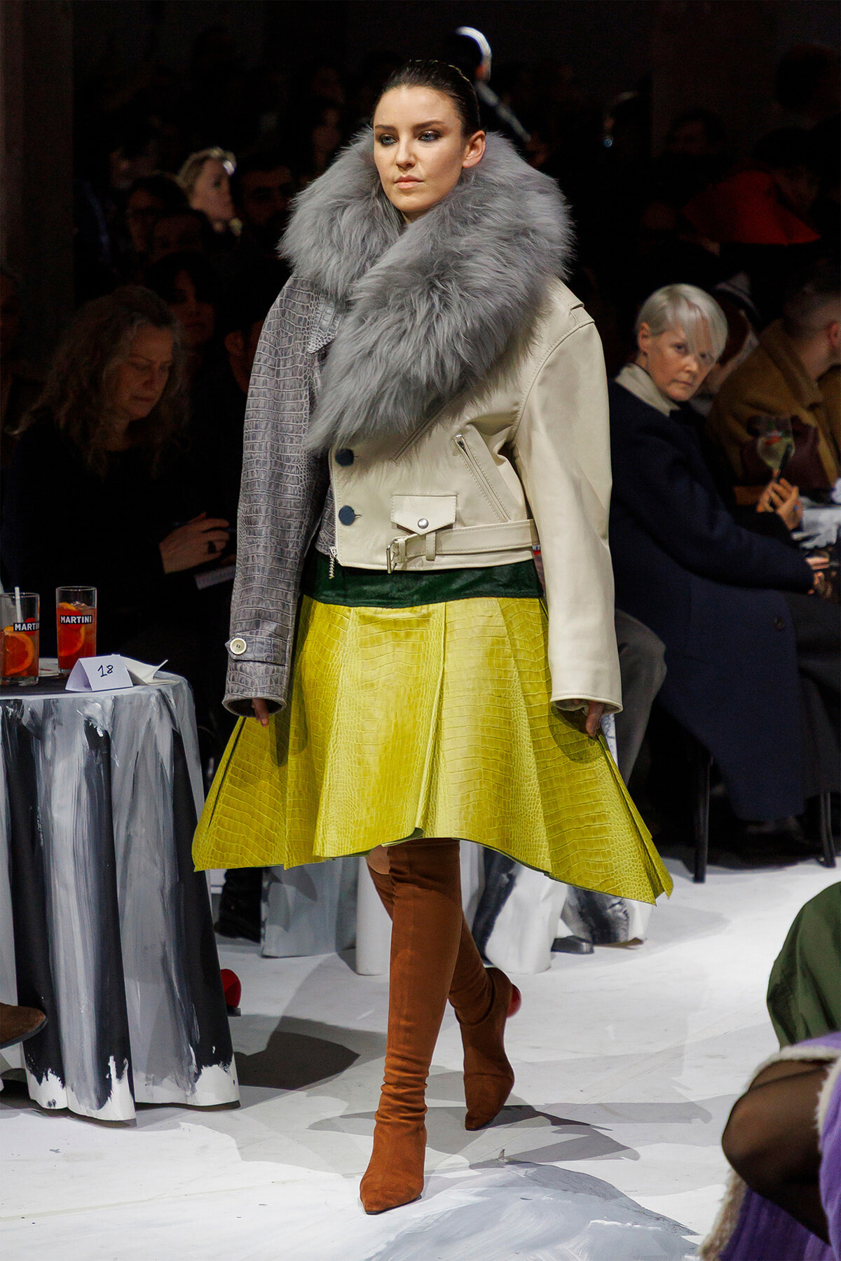 Model walks the runway in a cream leather jacket with an oversized gray fur collar, a bright yellow textured skirt, and brown suede thigh-high boots.