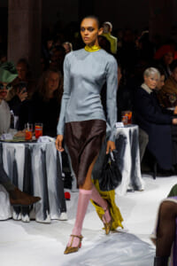 Model walking a runway in a pale blue long-sleeve top, brown slit-front skirt, pink tights, leopard-print heels, and a black handbag; audience in background.