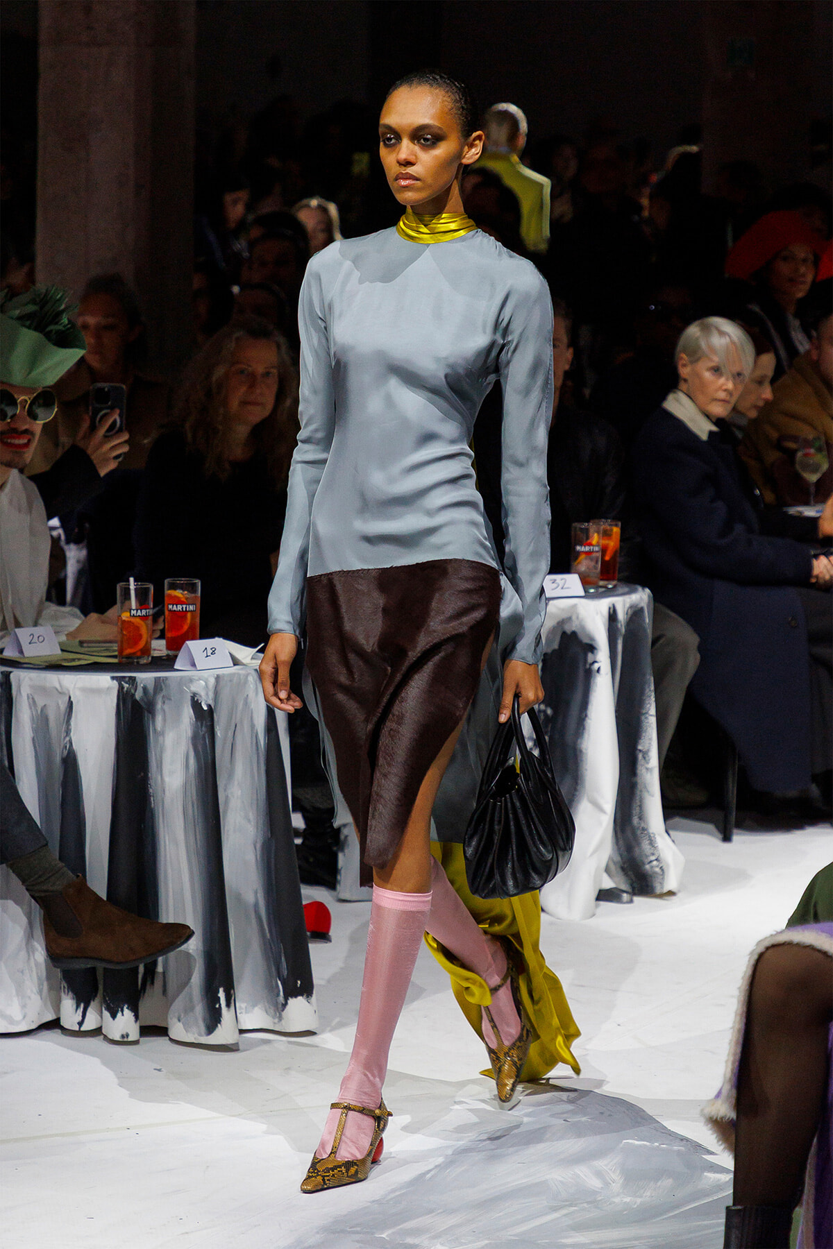 Model walking a runway in a pale blue long-sleeve top, brown slit-front skirt, pink tights, leopard-print heels, and a black handbag; audience in background.