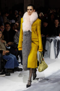 Model walks the runway in a bright yellow coat with a pink & gray fur scarf and pale yellow handbag amidst a seated audience.