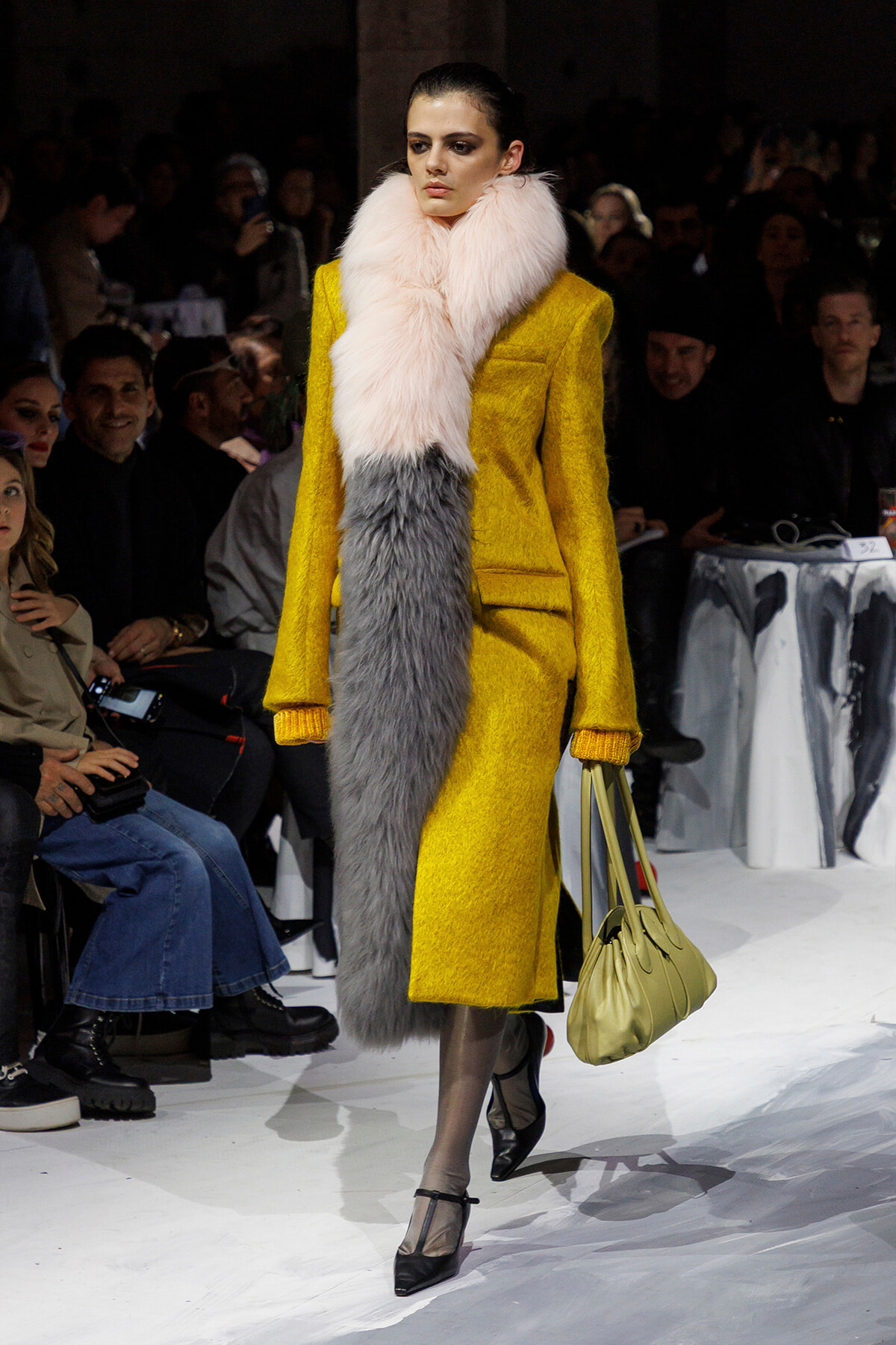 Model walks the runway in a bright yellow coat with a pink & gray fur scarf and pale yellow handbag amidst a seated audience.