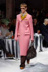Model on runway wearing a pink long coat with mustard lapel and pocket, brown pants, orange-tinted sunglasses, and carrying a dark brown handbag.