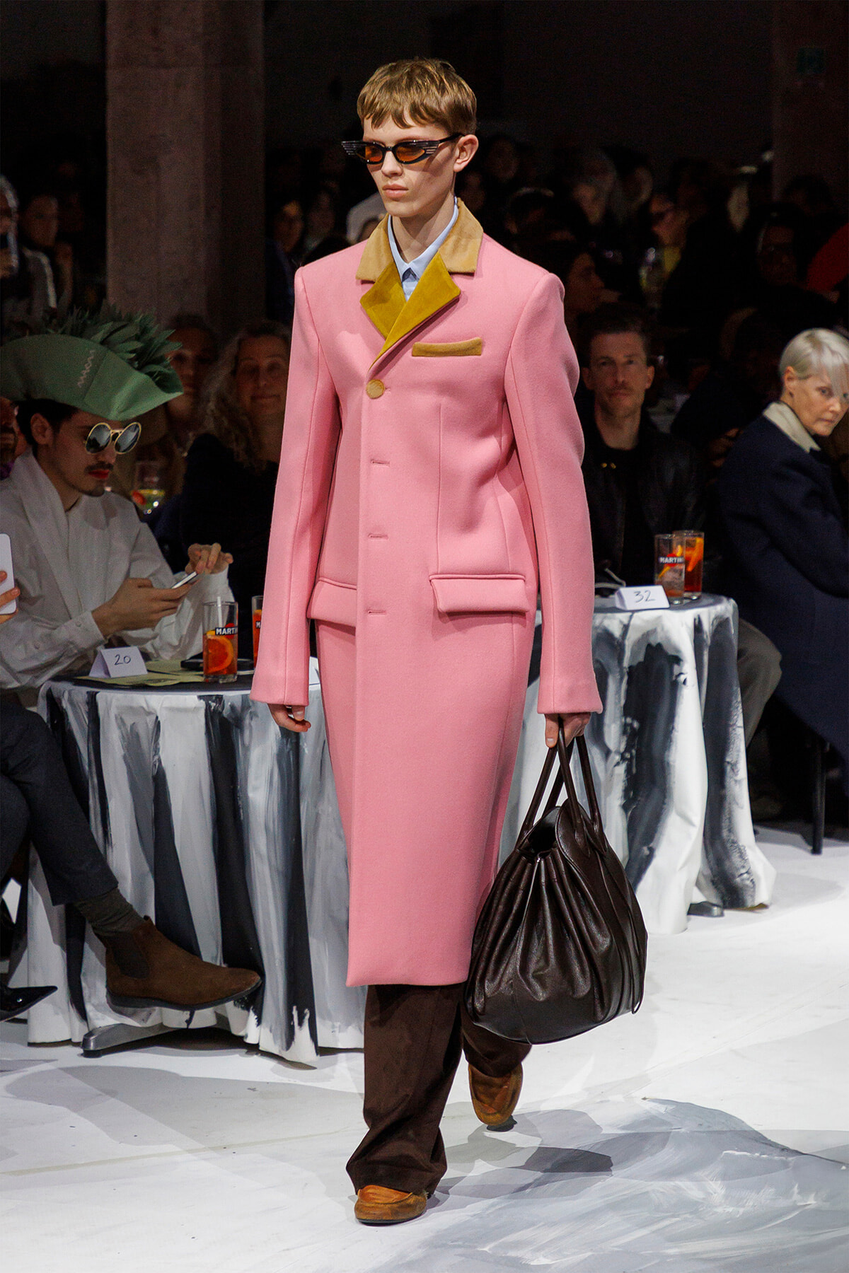 Model on runway wearing a pink long coat with mustard lapel and pocket, brown pants, orange-tinted sunglasses, and carrying a dark brown handbag.