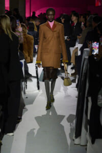 Model on runway wearing a camel coat over a pink turtleneck, yellow-cuffed sleeves, gray tights, yellow heels, and carrying a beige handbag amidst spectators.3a8🧥👠