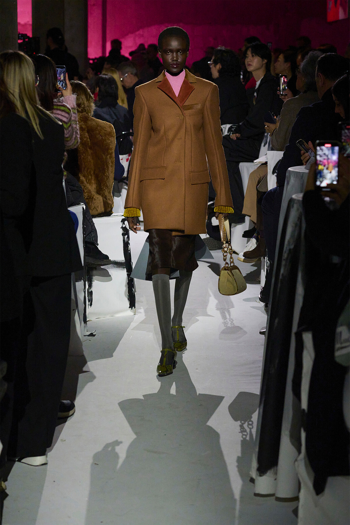 Model on runway wearing a camel coat over a pink turtleneck, yellow-cuffed sleeves, gray tights, yellow heels, and carrying a beige handbag amidst spectators.3a8🧥👠