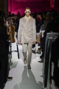 Model walking the runway in a beige double-breasted suit with yellow accents, white sunglasses, and snake print boots, pink-lit audience in background