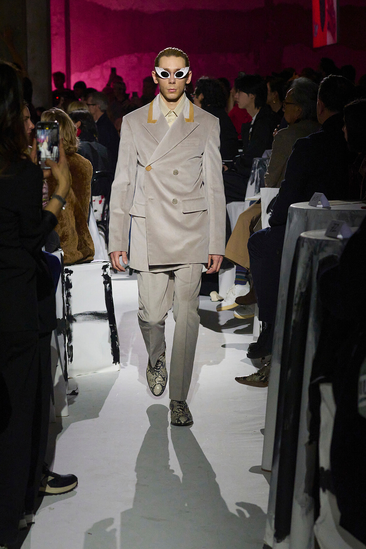Model walking the runway in a beige double-breasted suit with yellow accents, white sunglasses, and snake print boots, pink-lit audience in background