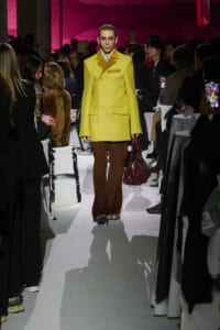 Model in a bright yellow double-breasted coat, brown flared pants, and burgundy handbag walks a white runway at a fashion show as the audience snaps photos on both sides.