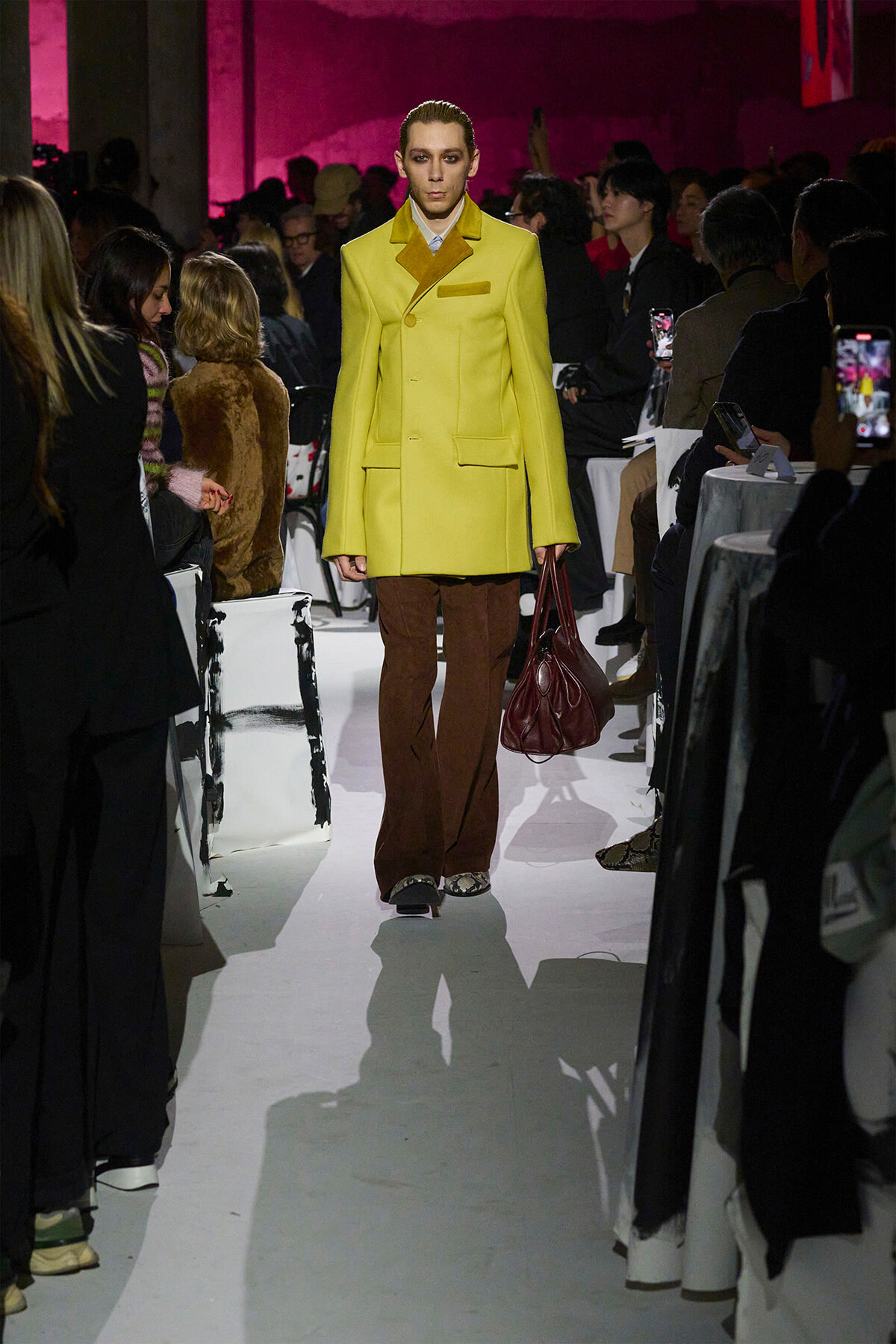 Model in a bright yellow double-breasted coat, brown flared pants, and burgundy handbag walks a white runway at a fashion show as the audience snaps photos on both sides.