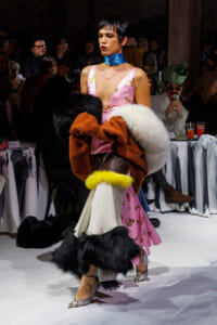 Model walks the runway in a pink butterfly-print dress with a bright blue neck scarf and a large fur stole draped over her arms as the audience watches.