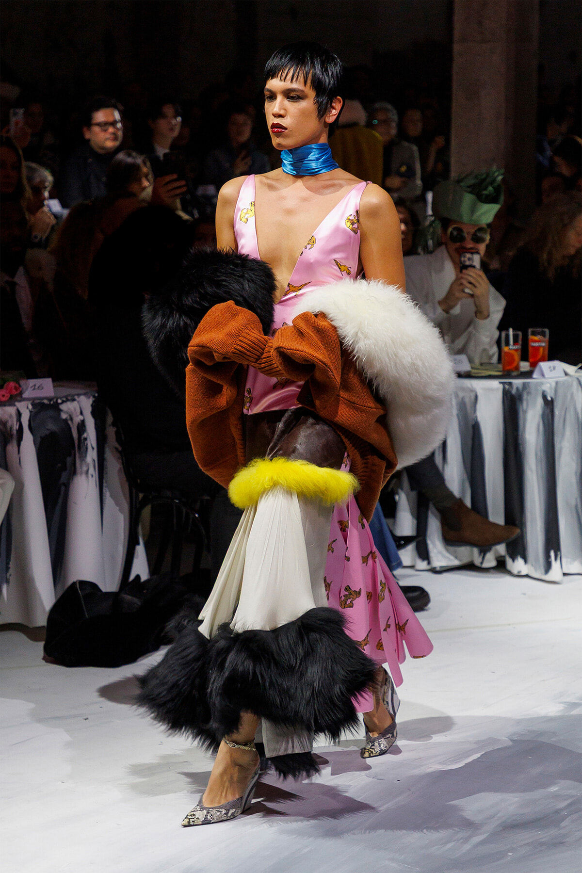 Model walks the runway in a pink butterfly-print dress with a bright blue neck scarf and a large fur stole draped over her arms as the audience watches.