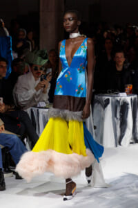 Model walks the runway in a cobalt blue satin V-neck top with floral embroidery and a bright yellow skirt with fur trim.