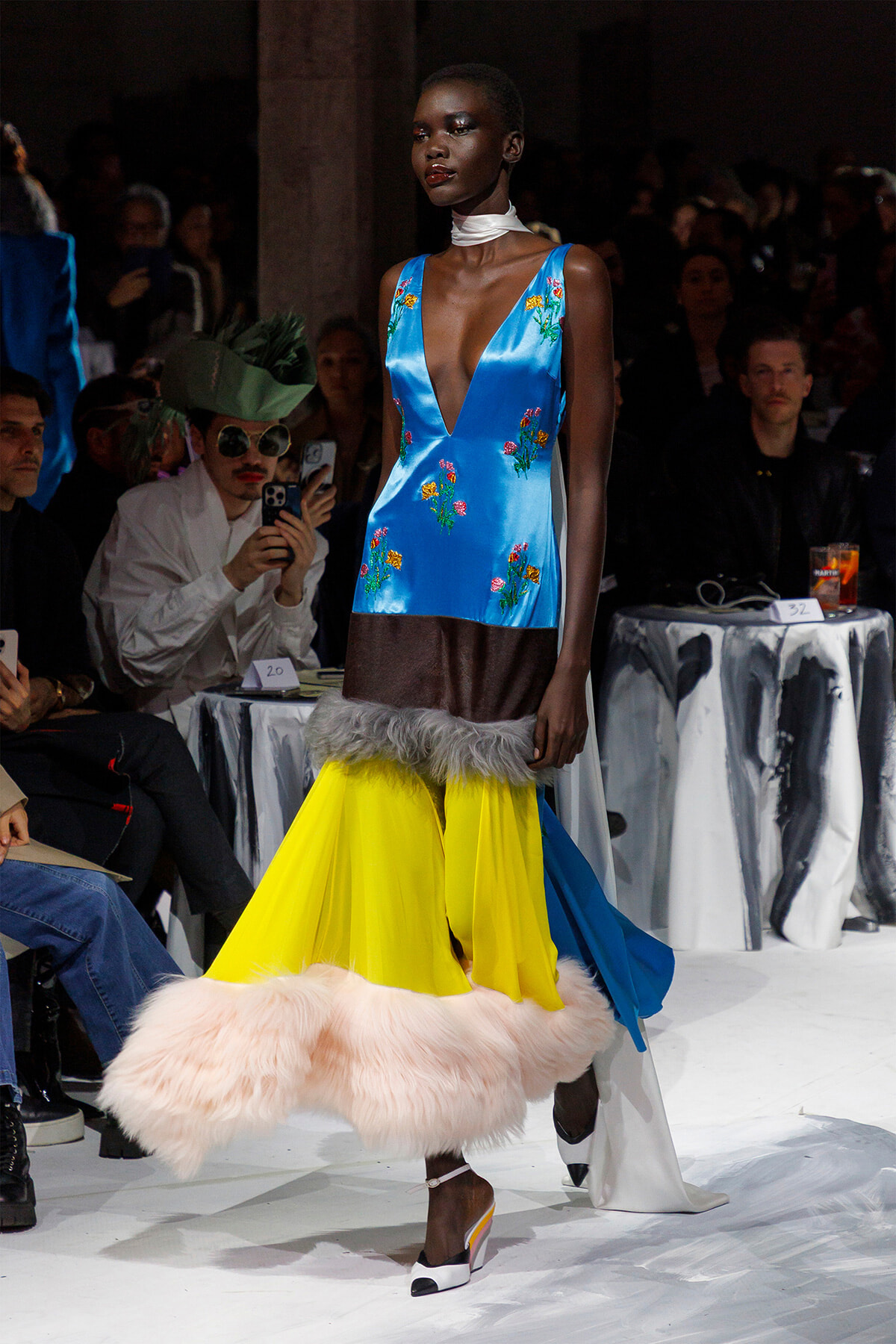 Model walks the runway in a cobalt blue satin V-neck top with floral embroidery and a bright yellow skirt with fur trim.