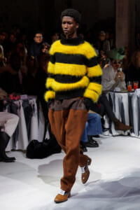 Model on a runway wearing a bright yellow and black fuzzy striped sweater with brown pants, walking the catwalk.