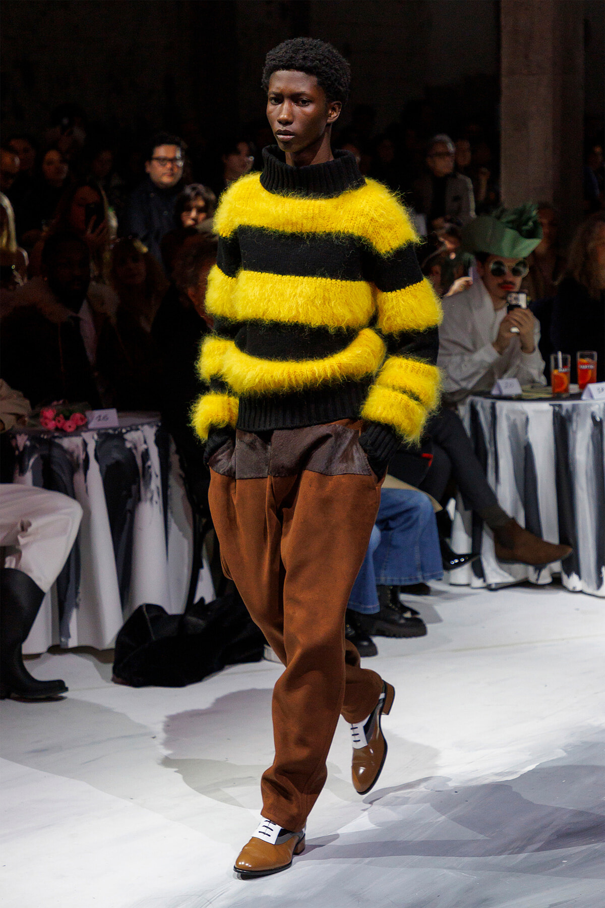 Model on a runway wearing a bright yellow and black fuzzy striped sweater with brown pants, walking the catwalk.