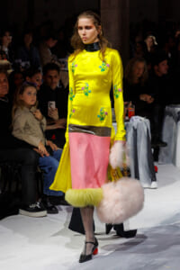 Model wearing a bright yellow dress with floral embroidery on the runway, paired with pink panel skirt and fluffy pink bag, walking past a seated audience.
