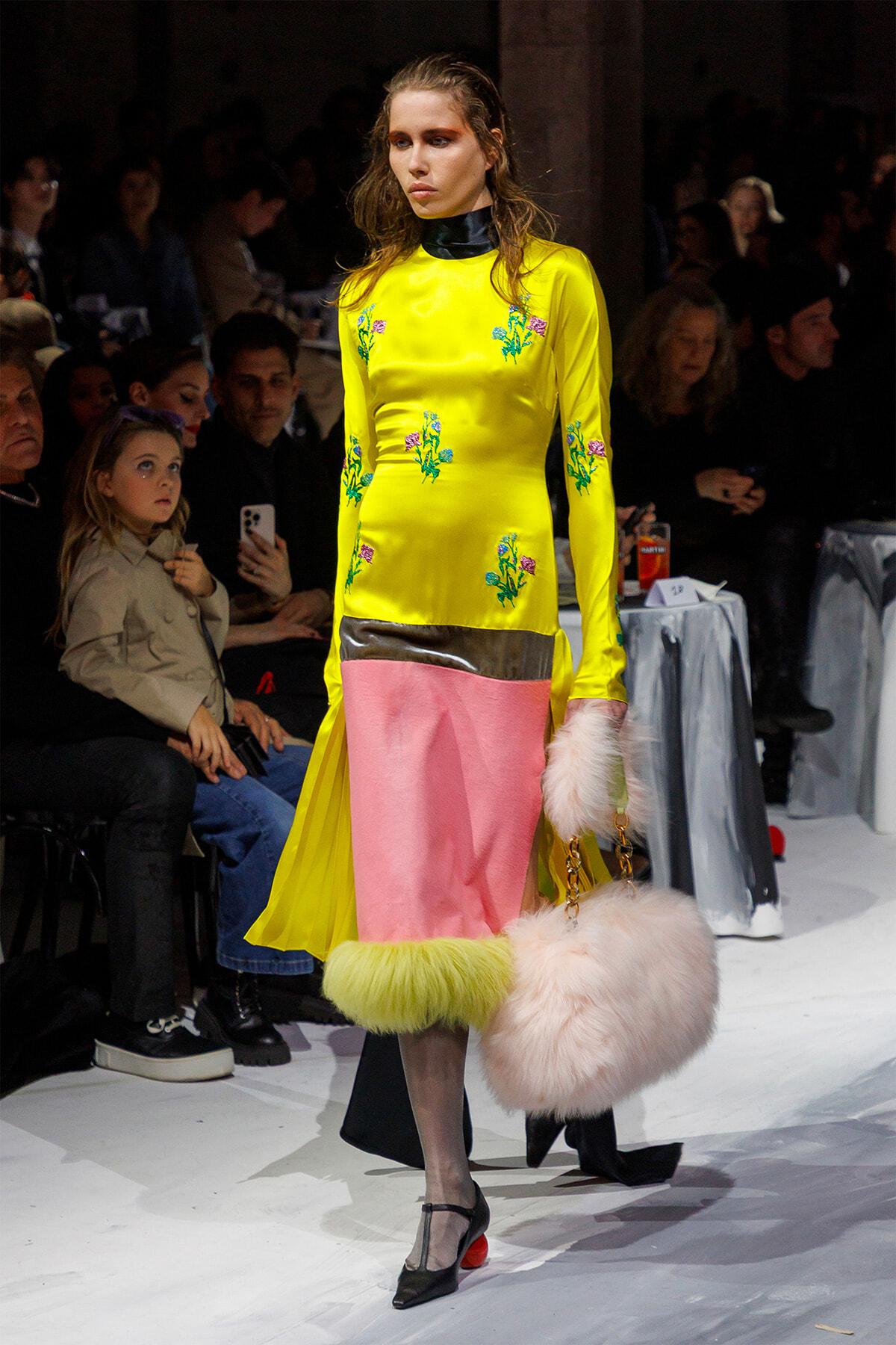 Model wearing a bright yellow dress with floral embroidery on the runway, paired with pink panel skirt and fluffy pink bag, walking past a seated audience.