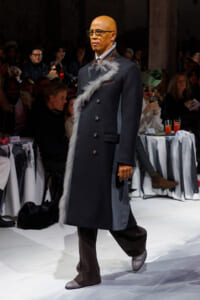Male model walks a runway in a dark double-breasted coat with gray fur trim, brown boots, and glasses, audience in the background.