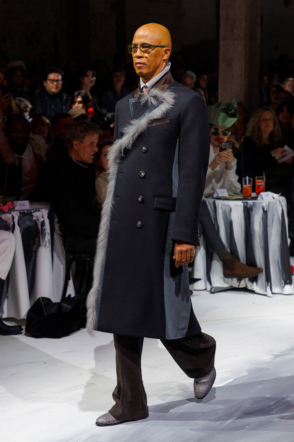 Male model walks a runway in a dark double-breasted coat with gray fur trim, brown boots, and glasses, audience in the background.