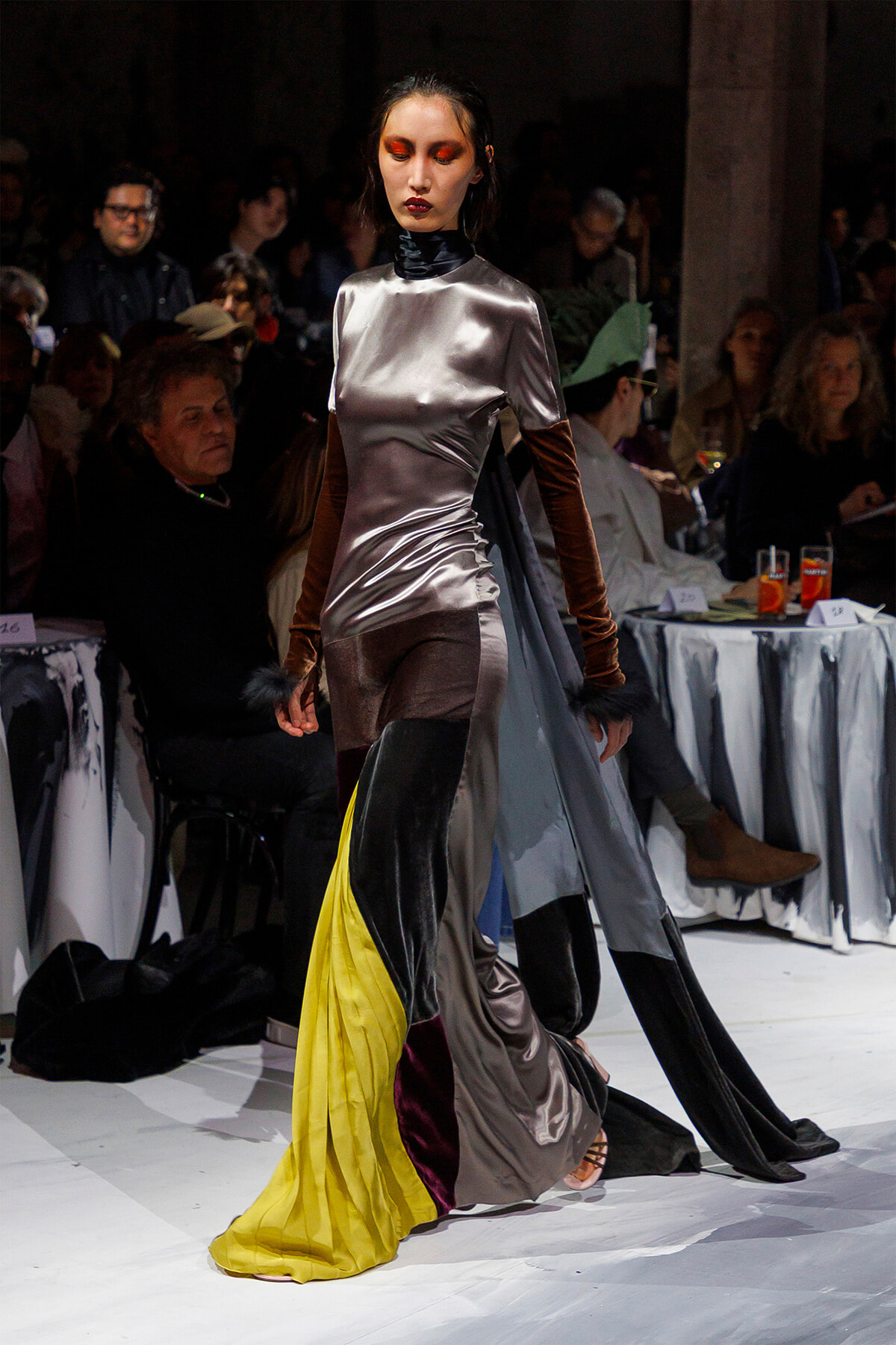 Model walks the runway in a silver satin gown with a colorful trailing train, audience visible in the background.