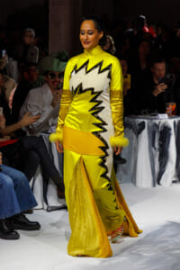 Model walks the runway in a neon yellow gown with jagged black zigzag trim and white panel, dramatic high neckline and feathered cuffs, surrounded by an attentive audience.