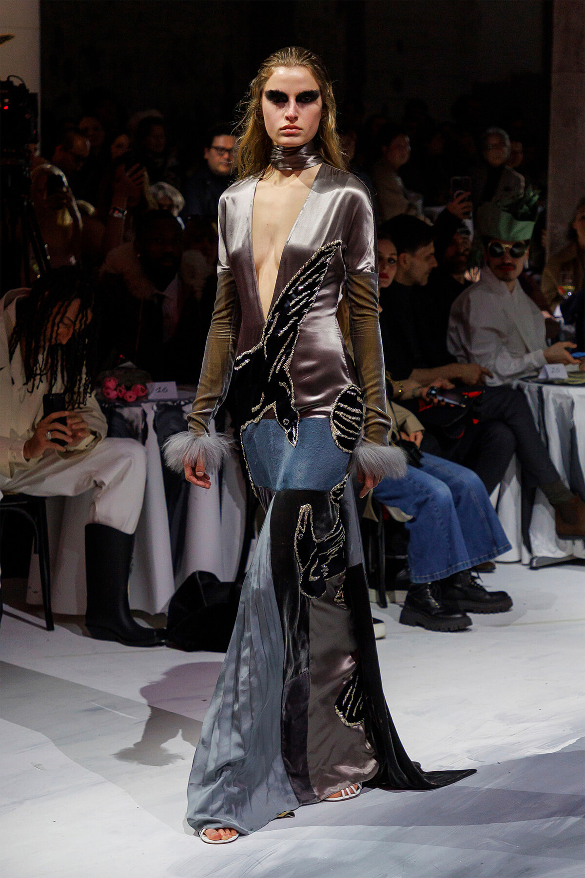 Model walks the runway in a metallic silver-gray gown with a deep plunging neckline, long sleeves, feathered cuffs, and black sequined appliqués on the skirt edge.