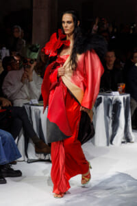 Model walking a fashion show in a red satin gown with a black feather shawl and voluminous pink-red cape.