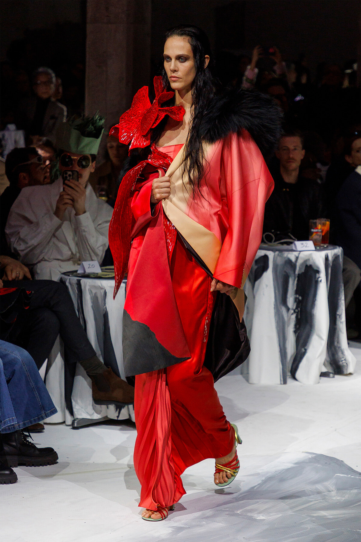 Model walking a fashion show in a red satin gown with a black feather shawl and voluminous pink-red cape.