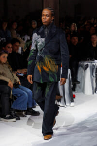 Male model walking a fashion runway in a dark floral-print suit with brown shoes, audience seated along the sides.