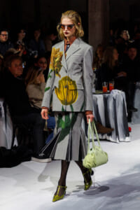 Model walks the runway in a gray floral-print blazer and matching skirt, with a pale green handbag and green heels, wearing sunglasses.