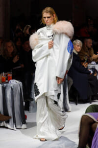 Model on runway wearing an oversized white coat with a pale pink fur collar and a blue patch, audience in background.