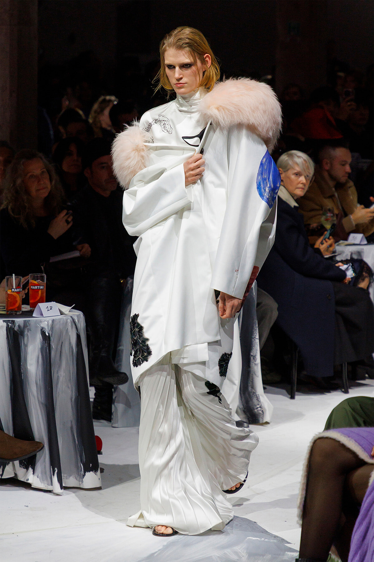 Model on runway wearing an oversized white coat with a pale pink fur collar and a blue patch, audience in background.
