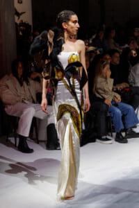Model in a white satin gown with gold embroidery and a black feathered sculptural accessory on the runway.