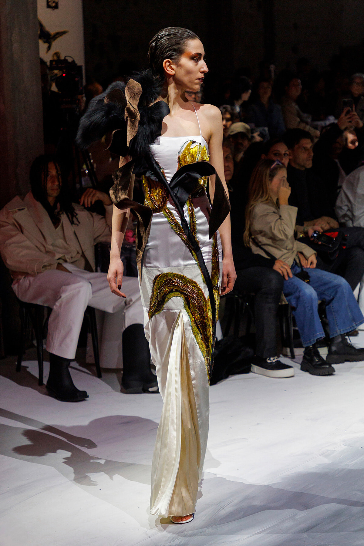 Model in a white satin gown with gold embroidery and a black feathered sculptural accessory on the runway.