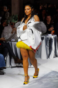 Model walks the runway wearing a white oversized coat with gray fur stole and bright yellow fur trim at the hem; yellow-and-black heels visible.