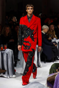 Male model walking a runway in a bold red suit with black textured embroidery resembling leaves or feathers across the jacket and pants, indoors at a fashion show.