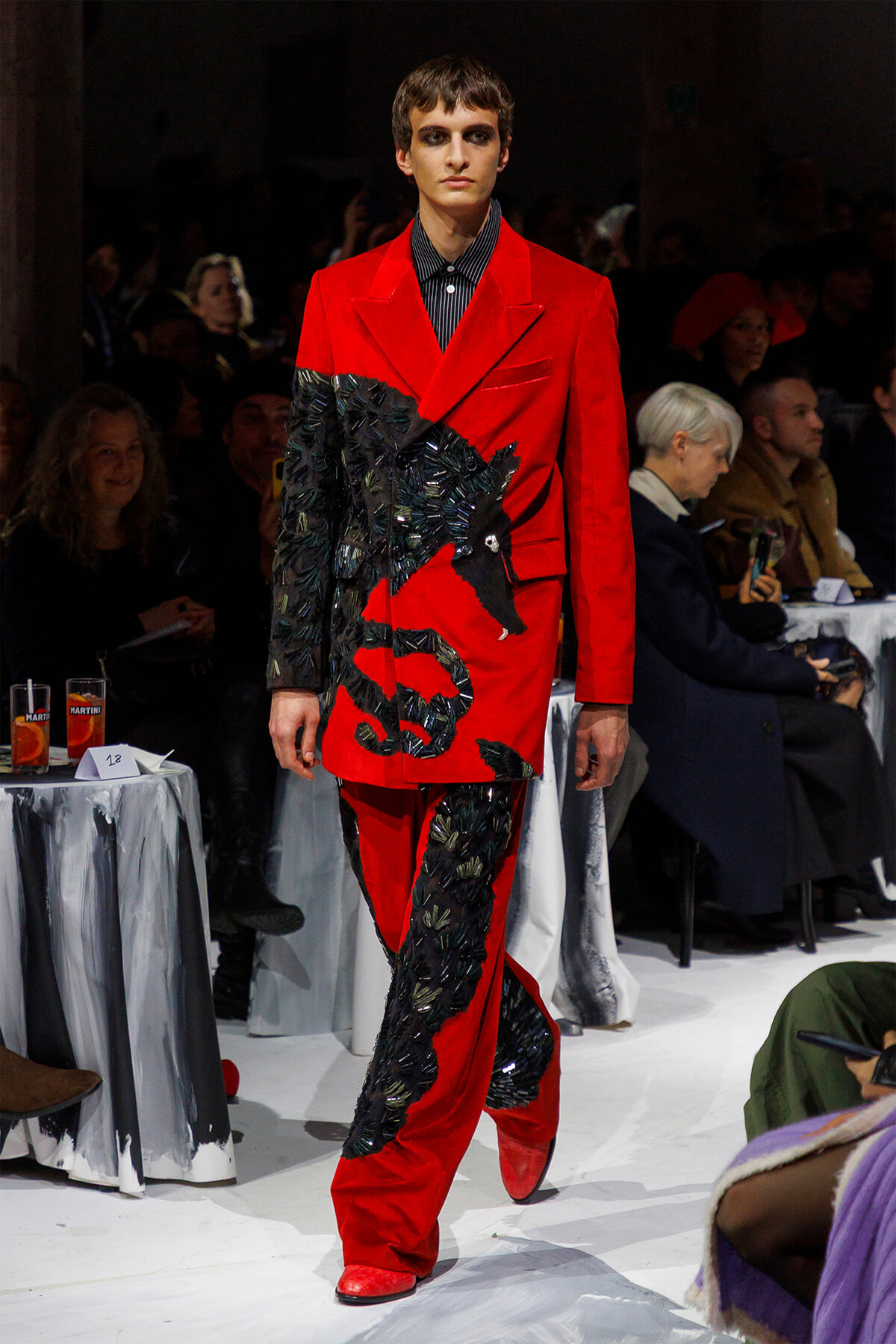 Male model walking a runway in a bold red suit with black textured embroidery resembling leaves or feathers across the jacket and pants, indoors at a fashion show.