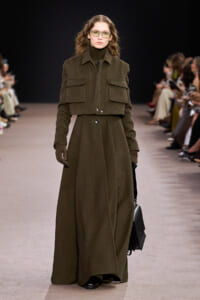 Model walks the runway in an oversized olive-green coat with large front pockets, matching gloves, and sunglasses, carrying a black bag.