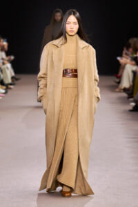 Model walks a runway in a full-length camel coat over a matching tan knit turtleneck and ribbed pants cinched with a wide brown belt.