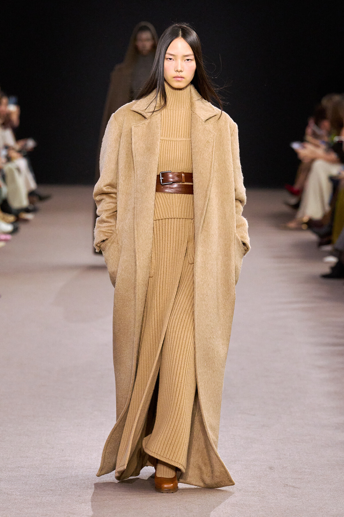 Model walks a runway in a full-length camel coat over a matching tan knit turtleneck and ribbed pants cinched with a wide brown belt.