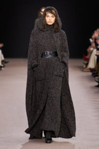 Model walking a runway in a long textured charcoal hooded cape, belted at the waist with pockets visible.