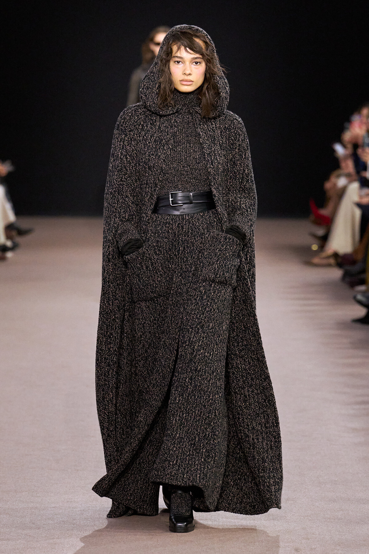 Model walking a runway in a long textured charcoal hooded cape, belted at the waist with pockets visible.