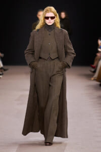 Model walking a runway in a brown tailored suit with a long overcoat and gloves, wearing dark sunglasses.