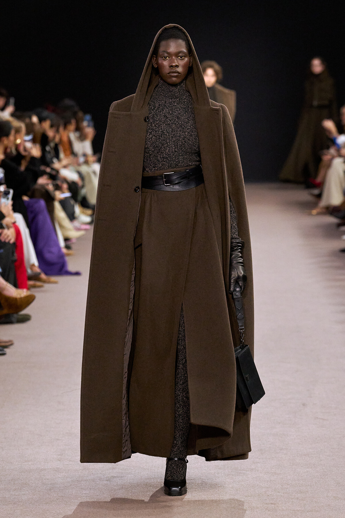 Model on a runway wearing a long brown hooded coat over a dark knit outfit with a wide black belt and black gloves.