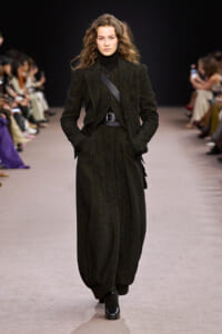 Runway model in a dark, textured suit and long coat, with a black turtleneck, wide belt, and a crossbody strap on a fashion show catwalk.
