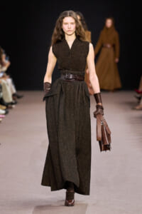 Model walking the runway in a dark brown sleeveless textured maxi dress with a wide belt, brown gloves, and a brown leather handbag.